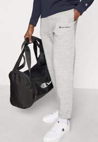 Γκρι φόρμα με ελαστικά μανσέτες, με λογότυπο σε ναυτικό μπλε. Μαύρη τσάντα duffel με λευκό λογότυπο κρατιέται από χέρι. Φοριούνται λευκά αθλητικά παπούτσια.