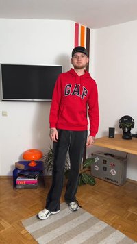 Camisola com capuz vermelha de algodão com logótipo "GAP", calças pretas e ténis prateados. Em pé sobre um tapete às riscas com uma televisão ao fundo.