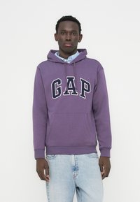 Sweat à capuche violet avec un grand logo "GAP" blanc, capuche à cordon, poche avant, porté par-dessus une chemise à col bleu clair, associé à un jean clair.