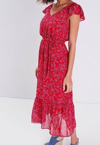 Robe maxi florale rouge avec de courtes manches volantées, une taille cintrée et une jupe à volants. Tissu léger avec un motif délicat.