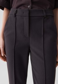 Pantalons taille haute gris foncé avec des plis, devant plat, passants de ceinture, poches latérales et une texture lisse. Design ajusté.