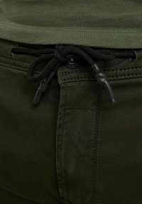 Olijfgroene broek met een trekkoord in de taille, voorzien van zwarte hardware, een gladde textuur en dubbel stiksel langs de naden.