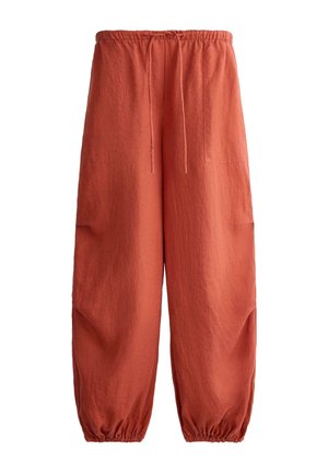 Nő fehér ujjatlan felsőt, laza rozsda színű, húzózsinóros nadrágot, fekete szandált és zöld fejkendőt visel, egyszínű háttér előtt áll.