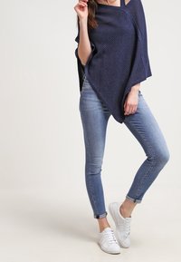 Marinblå stickad poncho med en strukturerad yta, i kombination med ljusblå tajta jeans och vita sneakers. Enkel, avslappnad outfitdesign.