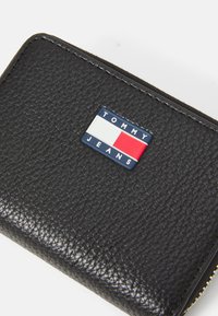 Billetera de cuero negro con textura de piedras y un parche del logo de Tommy Jeans en azul, rojo y blanco en la parte delantera.