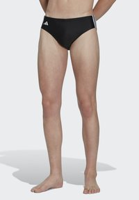 Des slip de bain noirs en tissu lisse avec trois bandes blanches sur la ceinture, conçus avec une taille basse pour un confort et une liberté de mouvement.