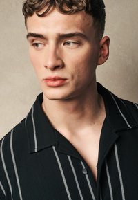 Camisa polo negra con relieve y rayas verticales blancas, que cuenta con un cierre de botones y un cuello con muesca, llevada por una persona con el cabello rizado y estilizado.