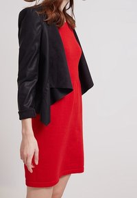 Giacca cropped in finta pelle nera su un vestito rosso aderente. La giacca presenta un colletto alto e maniche lunghe, mentre il vestito ha un collo rotondo.