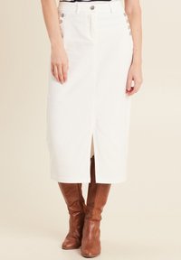 Jupe midi en denim blanc avec une fente à l'avant, ornée d'accents de boutons sur le côté. Portée avec des bottes marron jusqu'au genou.