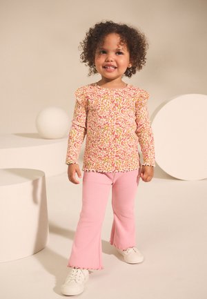 Top de flores de manga larga con volantes en los hombros, combinado con pantalones acampanados rosa con textura acanalada y zapatillas blancas. Fondo minimalista con formas suaves.