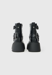 Botas de tobillo de cuero vegano negro con tacones de goma gruesos, correas ajustables y detalles del logo en la parte trasera. Acabado suave y diseño resistente.