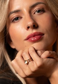 Zelta gredzens ar apaļu akmeni, ar gludu tekstūru un savienotiem akcentiem. Roka maigi balstās uz ādas, ar neitrālu fonu.