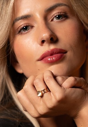 Gouden ring met een ronde steen, met een gladde textuur en gekoppelde accenten. Hand rust zachtjes tegen de huid, met een neutrale achtergrond.