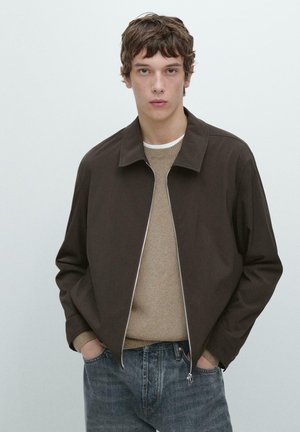 Giovane uomo che indossa una giacca marrone scuro con cerniera sopra un maglione beige e jeans blu, in piedi con le mani nelle tasche davanti a uno sfondo semplice.