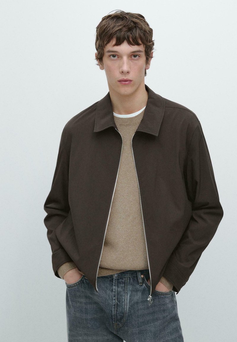 Jonge man draagt een donkerbruine ritssluitingjas over een beige trui en blauwe spijkerbroek, staand met handen in de zakken tegen een effen achtergrond.