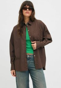 Camicia marrone a bottoni con taschino sul lato sinistro, indossata sopra un top verde aderente, abbinata a jeans in denim blu. Tessuto morbido con una vestibilità rilassata.