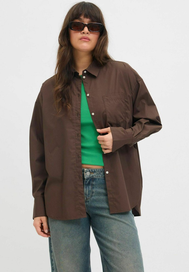 Camicia marrone a bottoni con taschino sul lato sinistro, indossata sopra un top verde aderente, abbinata a jeans in denim blu. Tessuto morbido con una vestibilità rilassata.