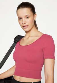 Tett, kortermet topp i ribbestruktur, rosafarge, med båtutringning og cropped lengde. Svart stropp fra en veske synlig.