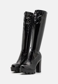 Mustat patenttikiiltonahasta valmistetut saappaat, joissa on paksu korko, pyöreä kärki ja sivuvetoketju. Käärmeenkäynnin teksturoitu pohja lisää korkeutta.