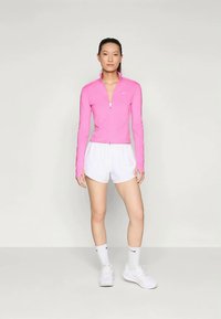 Giacca sportiva rosa aderente con zip e maniche lunghe, abbinata a pantaloni corti bianchi. Il modello indossa sneakers bianche e calzini alla caviglia.