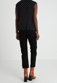 Une personne portant un haut sans manches à pois noirs, un pantalon noir et des bottines à talons marron, se tenant de dos sur un fond uni.