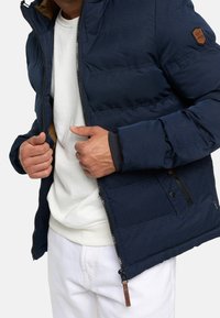 Granatowa puchowa kurtka z zamkiem błyskawicznym, z teksturowaną powierzchnią, brązową skórzaną naszywką z logo i elastycznymi mankietami. Noszona na białej bluzie.