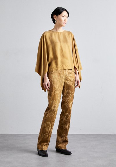Maglietta color senape con maniche ampie, abbinata a pantaloni marroni a fantasia con motivi floreali. Il modello si trova su un pavimento grigio.