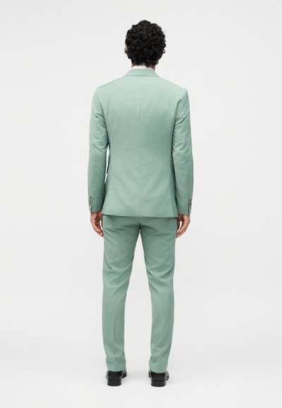 Άνδρας με σγουρά μαλλιά στέκεται με την πλάτη προς τα εμπρός, φορώντας ένα εφαρμοστό ανοιχτοπράσινο κοστούμι και μαύρα επίσημα παπούτσια σε ένα απλό λευκό φόντο.
