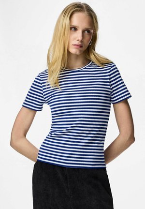 Lyshåret kvinde iført blå- og hvidstribet kortærmet skjorte og sorte bukser, stående med hænderne bag ryggen mod en hvid baggrund.