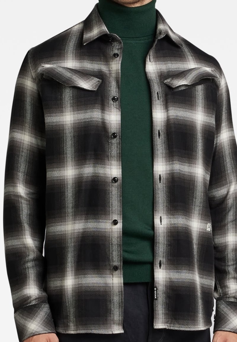 Homme portant une chemise à carreaux noir et blanc boutonnée sur un pull à col roulé vert foncé, montré des épaules aux hanches.