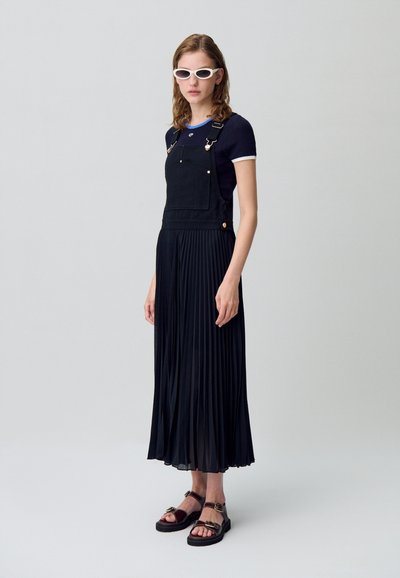 Salopette en denim noir avec une jupe plissée, un t-shirt côtelé bleu marine avec des bords blancs, et des sandales noires avec des accents en métal. Les accessoires incluent des lunettes de soleil blanches.