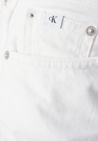 Bílá denimová tkanina má přední kapsu se stříbrnou hardware a malou značku s nápisem "cK". Texturovaný povrch s viditelnými detaily šití.