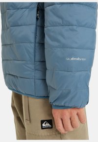 Ljusblå puffy-jacka med dragkedja och elastiska ärmslut. Har en diskret logotyp och quiltad struktur. Kompletteras av bruna byxor.