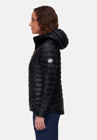 Kvinde i profil iført en sort quiltet hættetrøje med et hvidt logo på ærmet og marineblå bukser mod en hvid baggrund.