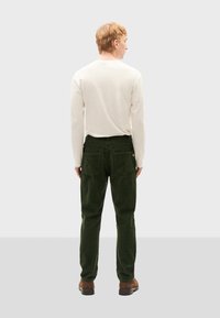 Grüne Cordhosen mit geradem Bein, ausgestattet mit Gesäßtaschen und einem Knopfakzent. Kombiniert mit einem langärmeligen weißen Hemd.