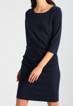 Mujer con un vestido azul marino ajustado de mangas tres cuartos, escote redondo y un leve fruncido en la cintura, de pie frente a un fondo liso.