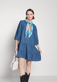 Robe en denim en forme de A avec des manches trois-quarts, des volants à plusieurs niveaux et une écharpe à motifs bleu clair. Bottes de cowboy noires et sac à main blanc.