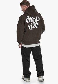 Dunkelbrauner Hoodie mit großem weißen "drop size"-Text auf dem Rücken. Hat eine lässige Passform, eine vordere Kängurutasche und gerippte Bündchen. Getragen mit schwarzen Hosen.