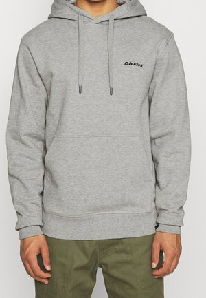 Person trägt eine hellgraue Kapuzenjacke mit "Dickies"-Logo auf der Brust, kombiniert mit olivgrünen Hosen, steht vor einem schlichten Hintergrund.