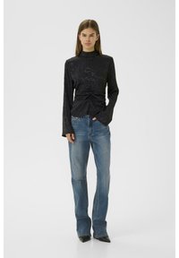 Haut noir à manches longues avec un motif floral texturé et un détail froncé à la taille, associé à un jean flared bleu clair et des chaussures noires à bout pointu.