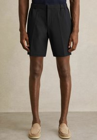Pantaloni scurți negri cu talie elastică, având un design frontal plat. Material neted și o croială ajustată, asortați cu pantofi slip-on de culoare deschisă.