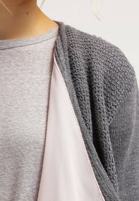 Cardigan grigio a maglia con un motivo testurizzato, indossato sopra una camicia grigia chiara e liscia con rifiniture rosa lungo il collo e le maniche.