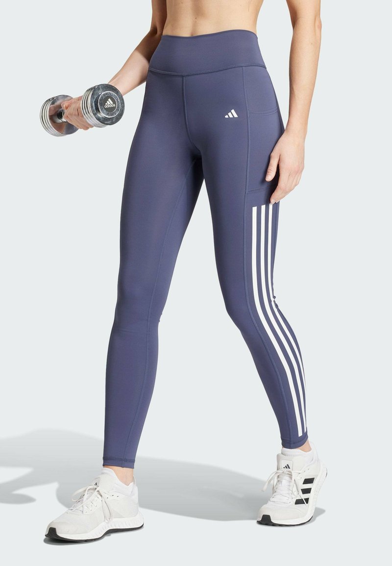 Navy hoge taille leggings met witte drie-strepen accenten aan de zijkant, gecombineerd met witte sportschoenen en een zilveren dumbbell.