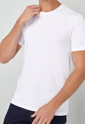 Homme portant un t-shirt blanc uni à manches courtes et col rond, et un pantalon foncé, ajustant le col de sa chemise avec la main droite.