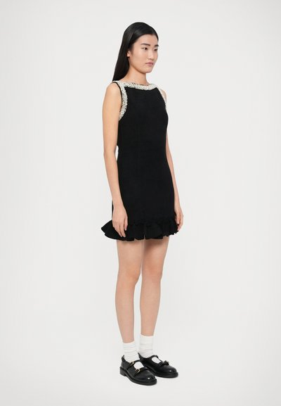 Svart ärmlös klänning med volangkant och kontrasterande pärlad halsringning; modellen bär svarta skor och vita strumpor.