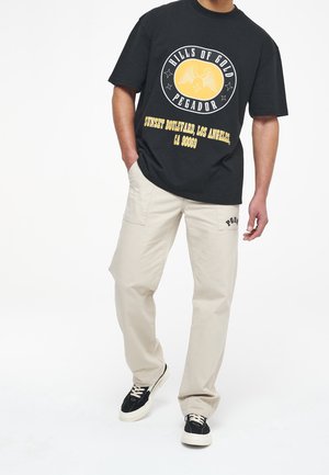 Hombre que lleva una camiseta gráfica negra con texto amarillo y pantalones beige, con zapatillas negras, de pie frente a un fondo blanco.