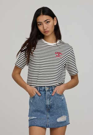 T-shirt a righe bianco e nere con collo rotondo e maniche corte, caratterizzato da testo ricamato rosso. Indossato con una gonna di jeans blu, leggermente strappata.
