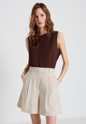 Donna con capelli mossi che indossa un top senza maniche marrone scuro e pantaloncini beige plissettati a vita alta con le mani nelle tasche.