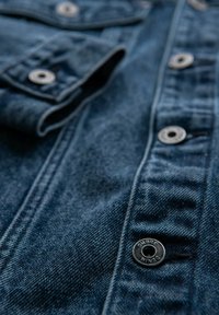 Camicia in denim blu scuro con bottoni argentati e cuciture visibili. Presenta un colletto e tasche con una superficie texturizzata per un tocco di dettaglio in più.