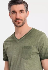 Olivově zelené tričko s výstřihem do V vyrobené z měkké látky, s přední kapsou na hrudi a ohrnutými krátkými rukávy pro uvolněný střih.